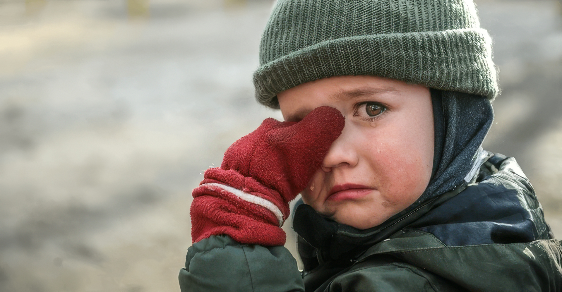 A young boy crying, wiping his eye with a red mitten